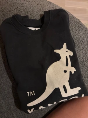 Svart sweatshirt från Kangol - Cool svart sweatshirt från Kangol med stor vit kängurulogga framtill. Tröjan har rund halsringning och ribbade muddar. Perfekt för en avslappnad streetwear-look.