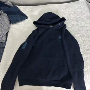 Mörkblå hoodie från USPA - Säljer en mörkblå hoodie från USPA med huva och snörning. Tröjan har en stor ficka framtill och ett tryckt USPA-logga på bröstet samt en siffra på ärmen. Perfekt för chill dagar och streetwear-stil.