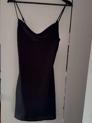 Svart satinklänning 32 GinaTricot  - Svart, kort klänning i satin ifrån Gina Tricot. Tunna, justerbara axelband och draperad urringning. Storlek 32, normal i storleken. Använd en gång. Se sista bilden för mått. 