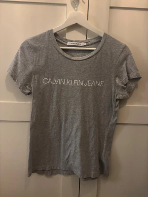 Grå Calvin Klein Jeans t-shirt - Snygg grå t-shirt från Calvin Klein Jeans med vit logga tryckt framtill. Klassisk rund halsringning och korta ärmar. Tillverkad i mjukt och bekvämt material som passar perfekt till jeans eller shorts.