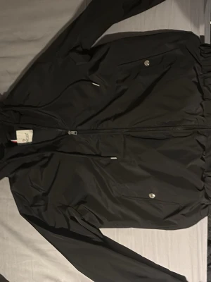 Svart vindjacka från Moncler - Snygg svart vindjacka från Moncler med huva och dragkedja framtill. Jackan har två fickor med tryckknappar, Moncler-logga på bröstet och detaljer i rött, vitt och blått på huvan. Tillverkad i ett lätt och vattenavvisande material.