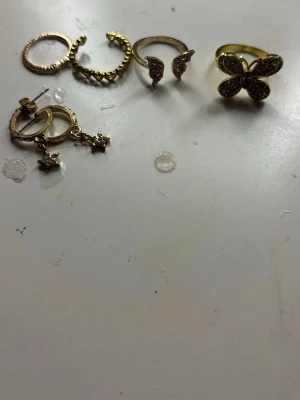 Mixade guldfärgade ringar och örhängen - Säljer ett set med guldfärgade smycken: flera ringar i olika former, bland annat en med fjäril och en med hjärtan, samt örhängen med små stjärnhängen. Alla smycken har dekorativa detaljer och gnistrande stenar för extra bling.