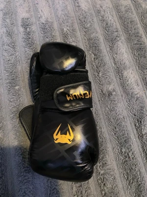Svarta boxningshandskar från Venum - Säljer ett par svarta boxningshandskar från Venum med guldfärgade loggor och detaljer. Handskarna har kardborreband för enkel justering och sitter skönt på handen. Perfekta för träning eller sparring inom kampsport.
