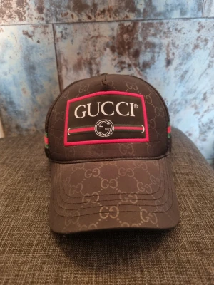 Brun Gucci keps med logotyp - Säljer en brun keps från Gucci med klassiskt GG-mönster över hela kepsen. Stor broderad logotyp framtill med röd och grön detalj samt svart bakgrund. Kepsen har böjd skärm och justerbar rem bak. Perfekt för dig som vill ha en lyxig och trendig accessoar.