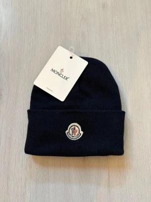 Mörkblå Moncler mössa med logga - Snygg mörkblå mössa från Moncler med klassisk uppvikt kant och Monclers ikoniska logotyp framtill. Tillverkad i mjukt stickat material som håller dig varm och stilren. Perfekt accessoar för kalla dagar och streetwear-vibe.