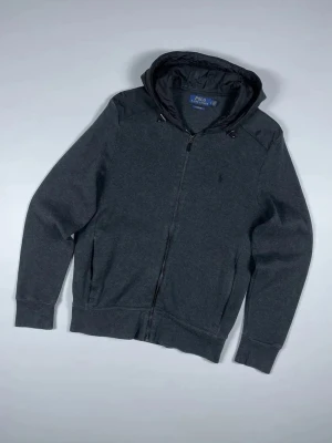 Ralph Lauren Cardigan  - Mörkgrå cardiganjacka med dragkedja från Polo Ralph Lauren helt utan defekter. Jackan har huva i glansigt material och matchande axelparti, samt ribbade muddar vid ärmslut och midja, perfekt nu när våren närmar sig. Klassisk och stilren design med diskret logga på bröstet. Kontakta mig om frågor!😊