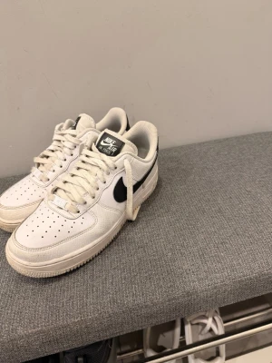 Nike Air Force 1 vita sneakers - Klassiska vita Nike Air Force 1 sneakers med svart swoosh på sidan. Skorna har perforerad tå för ventilation, vita skosnören och en robust sula. Tillverkade i läder med textildetaljer och låg profil som ger en clean och tidlös look.