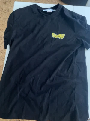 Svart Off-White t-shirt med tryck - Svart t-shirt från Off-White med färgglada tryck och loggor på ryggen samt ett mindre tryck på bröstet. T-shirten har korta ärmar och är i mjuk bomull med en klassisk passform och rund hals.