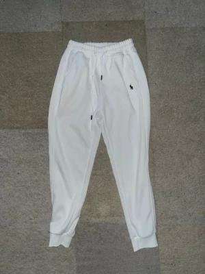 Vita mjukisbyxor från Polo Ralph Lauren - Säljer ett par fräscha vita mjukisbyxor från Polo Ralph Lauren med broderad logga på vänster sida. Byxorna har resår i midjan med dragsko, muddar vid bensluten och två sidofickor. Perfekta för en chill och stilren look.