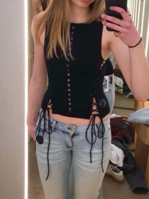 Svart korsettopp med snörning - Snygg svart korsettopp med snörning i sidorna och dekorativa knappar framtill. Toppen är ärmlös och har en croppad passform som ger en edgy vibe. Perfekt för dig som vill sticka ut och skapa en cool look. Köpt för 500kr och i nyskick💕