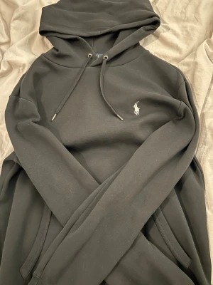 Svart hoodie från Polo Ralph Lauren - Svart hoodie från Polo Ralph Lauren med klassisk broderad logga på bröstet. Hoodien har dragsko i huvan och känguruficka framtill. Tillverkad i mjuk bomullsblandning som är skön att ha på sig. Perfekt för en avslappnad och stilren look.