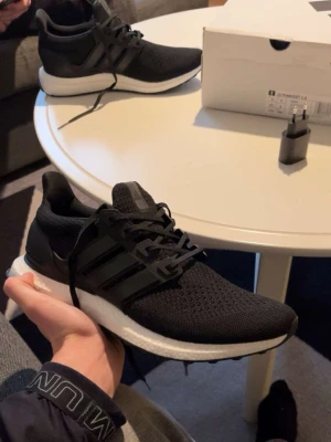 Adidas Ultraboost svarta sneakers - Svarta Adidas Ultraboost sneakers med stickad ovandel och klassiska tre ränder på sidan. Skorna har snörning och en vit, responsiv Boost-sula som ger grym komfort. Perfekta för dig som gillar stilrena och sportiga skor med modern känsla.