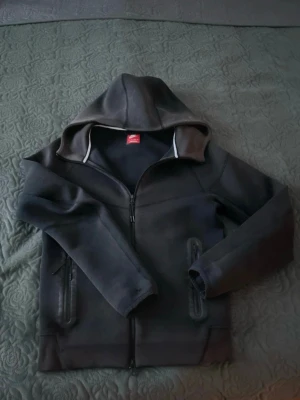 Svart Nike tech med dragkedja - Svart hoodie med hel dragkedja framtill och två fickor med dragkedja. Utrustad med huva och diskreta detaljer i grått vid kragen. Perfekt för en avslappnad och sportig stil.