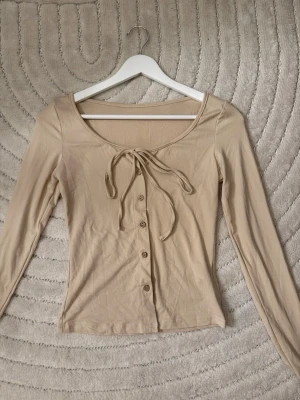 Beige långärmad topp med knyt - Trendig beige topp med långa ärmar, knappar framtill och snygg knytning vid halsringningen. Tillverkad i mjukt bomullsmaterial och har en figurnära passform som passar perfekt till jeans eller kostymbyxor. 