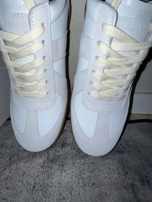Maison Margiela Replica vita sneakers - Maison Margiela Replica sneakers i vitt läder och mocka med beige snörning och klassisk gummisula. Snygg retrostil med diskreta detaljer och stilren design. Perfekta för dig som gillar minimalistiska och tidlösa sneakers.