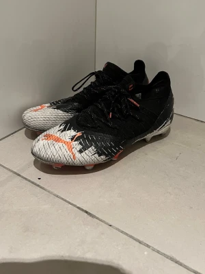 Puma svarta och vita fotbollsskor - Säljer ett par snygga fotbollsskor från Puma i svart och vitt med orange detaljer. Skorna har låg profil, snörning och mönstrad ovandel i syntetmaterial för bra grepp på planen. Perfekta för dig som vill sticka ut på matchen!