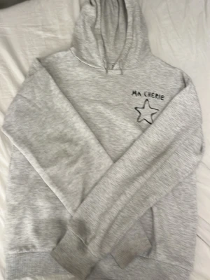 Grå hoodie med tryck 'Ma Chérie' - Ljusgrå hoodie med huva och trycket 'Ma Chérie' samt en svart stjärna på bröstet. Mjuk och skön bomullsblandning, ribbade muddar vid ärmslut och nederkant. Perfekt för en chill och avslappnad stil.