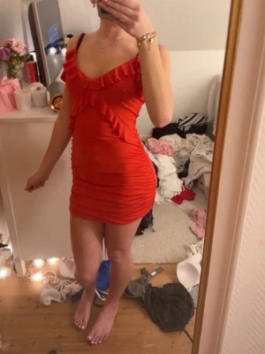 Röd volangklänning med v-ringning - Snygg röd kortklänning med volangdetaljer och v-ringad hals. Klänningen är figurnära och har smala axelband. Perfekt för dig som vill sticka ut med en färgstark och trendig look.