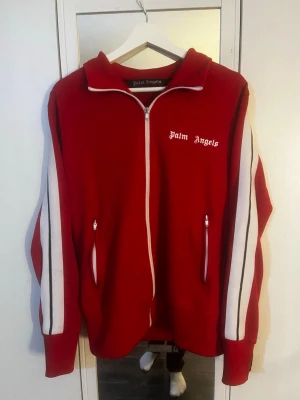 Röd zip hoodie från Palm Angels - Snygg röd hoodie från Palm Angels med vit dragkedja och vita ränder längs ärmarna. Loggan är broderad i vitt på bröstet. Hoodien har två fickor med dragkedja och sportig look. Tillverkad i mjukt material, perfekt för en avslappnad streetwear-stil. Stygnen har släppt lite på insidan annat helt perfekt. Hör av er vid frågor!