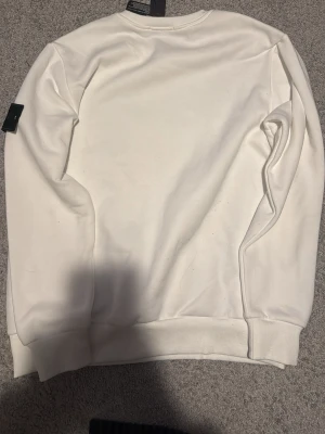 Vit sweatshirt från Stone Island - Säljer en clean vit sweatshirt från Stone Island med den klassiska svarta logotypen på vänster ärm. Tröjan har rund halsringning, ribbade muddar och är tillverkad i mjukt bomullsmaterial. Perfekt för en stilren och avslappnad look.