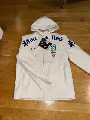 Vit Brasil Nike vindjacka med huva - Vit vindjacka från Nike med huva och dragkedja. Jackan har Brasil-logga, grönt Nike-märke och flera sponsortryck i blått och rött, inklusive Itaú och Guaraná. Tillverkad i GORE-TEX-material för extra skydd mot vind och regn. Perfekt för dig som gillar sportig stil. Står att den är stolek M sitter mer som S