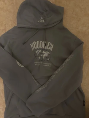 Grå hoodie från Hoodrich  - Snygg grå hoodie från Hoodrich med stort tryck på bröstet och detaljer på ärmarna. Klassisk huva med dragsko och magficka framtill. Hoodien har lång ärm och relaxed passform, perfekt för en streetwear-look. Den är använd 3 gånger bara 