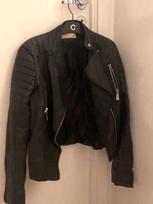 Svart biker skinnjacka med dragkedjor - Snygg svart skinnjacka i bikerstil med markerade axlar och detaljerade sömmar. Jackan har flera silvriga dragkedjor framtill och på ärmarna, samt en klassisk krage. Perfekt för en cool och trendig look.