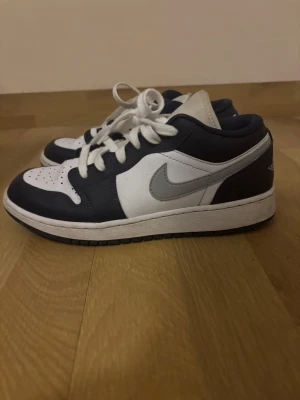 Nike Air Jordan 1 Low blå/vit - Snygga Nike Air Jordan 1 Low sneakers i blått och vitt med grå Swoosh. Klassisk siluett med lågt skaft, vita skosnören och perforerad tåbox. Ovandelen är i läder och yttersulan är svart. Perfekta för dig som gillar streetwear och ikoniska sneakers.