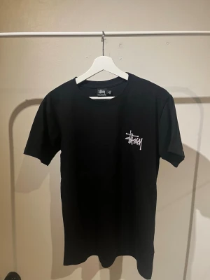 Svart Stüssy t-shirt med logga - Svart t-shirt från Stüssy med klassisk vit logga tryckt på bröstet. T-shirten har rund halsringning och korta ärmar, perfekt för en streetwear-look. Tillverkad i mjuk bomull för skön komfort.