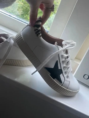 Vita sneakers med svart stjärna - Snygga vita sneakers med svart stjärndetalj på sidan och zebramönstrad häl. Skorna har vita snören och en beige sula med räfflad kant. Perfekta för dig som vill sticka ut lite extra med coola detaljer. Materialet ser ut att vara konstläder.