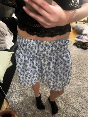 Vit och blå kortkjol med mönster - Kortkjol med smock i midjan och blått mönster på vit botten. Kjolen har en luftig och lätt form, perfekt för varmare dagar. Materialet känns mjukt och bekvämt mot huden. Den är runt storlek 146