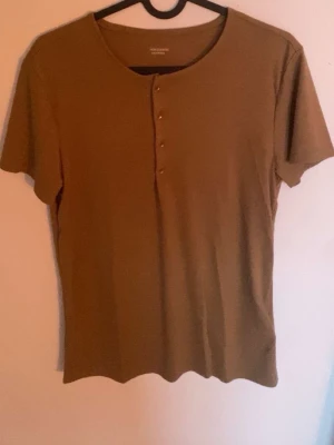 T-shirt med knappar - En enkel brun t-shirt med rund halsringning och korta ärmar. Tröjan har en rak passform och fyra knappar framtill som detalj. Perfekt att styla med jeans eller linnebyxor för en clean look. Köpt på Franska rivieran. 