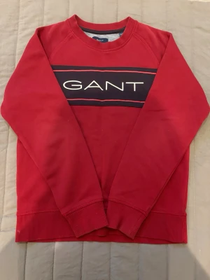 Röd sweatshirt från GANT - Röd sweatshirt från GANT med stort logotryck över bröstet i vitt på en mörkblå panel. Tröjan har rund halsringning, ribbade muddar vid ärmslut och nederkant samt en klassisk, avslappnad passform. Perfekt för en casual look.
