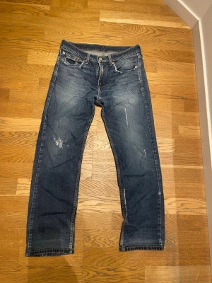 Levi's 514 - Säljer ett par klassiska blå Levi's jeans med raka ben och slitningar på både fram- och baksida. Jeansen har fem fickor och den ikoniska röda Levi's-lappen bak. Tillverkade i slitstark denim, perfekta för en avslappnad streetstil. Jeansen är i längd 32 men uppsydda till 30
