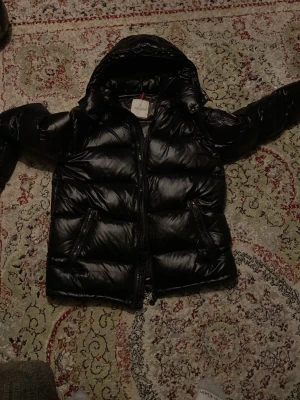 Svart dunjacka från Moncler - Snygg svart dunjacka från Moncler med glansig finish och huva. Jackan har två dragkedjeförsedda fickor och Moncler-logga på ärmen. Perfekt för kalla vinterdagar och riktigt trendig look.