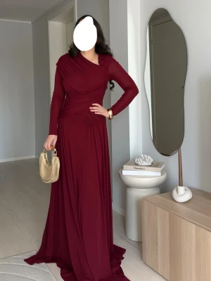 Lång vinröd klänning med drapering - Elegant vinröd långklänning med draperade detaljer och långärmad design. Klänningen har en omlottliknande överdel och markerad midja som ger en smickrande siluett. Tillverkad i ett mjukt, flytande material som faller snyggt längs kroppen.