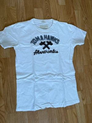 Vit t-shirt Abercrombie  - Vit t-shirt från Abercrombie & Fitch med broderad text 'Tomahawks' och två korsade yxor på bröstet. Klassisk rund hals och korta ärmar. Perfekt för en avslappnad stil.
