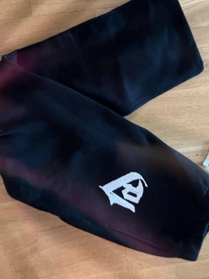 Svarta mjukisbyxor med vit logga - Säljer ett par svarta mjukisbyxor med en stor vit broderad logga på ena benet. Byxorna har en avslappnad passform och är gjorda i mjukt material, perfekta för chill dagar. Resår i midjan och vid benslut för extra komfort.