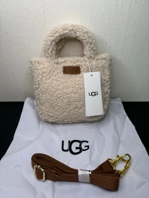 Beige teddy handväska från UGG - Supersöt beige handväska från UGG i mjukt teddy-material. Väskan har korta handtag, avtagbar axelrem i brunt tyg med guldfärgade detaljer och UGG-logga framtill. Perfekt för dig som gillar mysig och trendig stil.