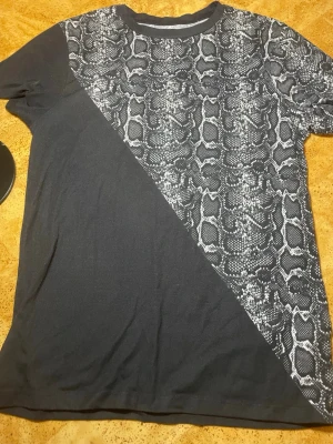 Svart t-shirt med ormmönster - Säljer en svart t-shirt med diagonal ormmönstrad panel i vitt och grått. T-shirten har korta ärmar och är gjord i mjuk bomull. Perfekt för dig som vill sticka ut med ett unikt mönster.