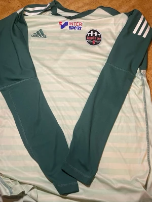 Grön långärmad träningströja Adidas - Säljer en grön långärmad träningströja från Adidas med vita ränder på axeln och diskreta ljusgröna ränder på kroppen. Tröjan har tryck från Intersport och Årsta FF på bröstet. Perfekt för fotbollsträning eller uppvärmning.