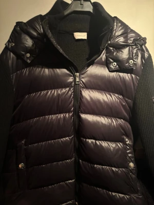 Svart dunväst från Moncler - Snygg svart dunväst från Moncler med glansig finish och quiltad design. Västen har dragkedja framtill, två fickor med tryckknappar och hög krage. Perfekt för lager på lager och ger en clean look till din outfit.