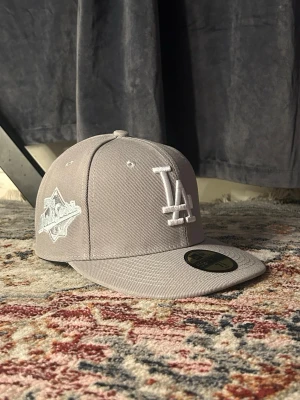 Grå LA Dodgers keps från New Era - Snygg grå keps från New Era med LA Dodgers-logga broderad i vitt framtill och World Series 1988-märke på sidan. Klassisk flat brim och justerbar passform. Perfekt för dig som gillar sportig streetstyle och vill ha en stilren accessoar.