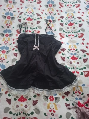 Svart nattlinne med spets och rosett - Ett svart nattlinne med tunna axelband, dekorerat med vit spets längs nederkanten och en söt ljusrosa rosett framtill. Nattlinnet har en lätt utsvängd form och detaljer med spets längs bysten. Perfekt för en mysig kväll hemma.