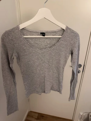 Grå långärmad topp med spetskant - Säljer en enkel och stilren grå långärmad topp i mjuk bomullsmix. Toppen har en rundad halsringning med fin spetskant som ger en söt detalj. Perfekt att matcha med jeans eller kjol för en avslappnad look.