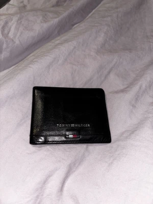 Svart plånbok Tommy Hilfiger - Snygg svart plånbok från Tommy Hilfiger i äkta läder. Klassisk design med diskret logga och en liten detalj i Hilfigers färger på framsidan. Perfekt storlek för kort och sedlar, stilren och enkel att ha med sig. Priset kan diskuteras