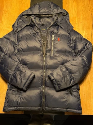 Mörkblå dunjacka från Polo Ralph Lauren - Snygg mörkblå dunjacka från Polo Ralph Lauren med huva och dragkedja framtill. Jackan har en bröstficka med dragkedja och den klassiska röda loggan på bröstet. Puffig modell som håller dig varm under vintern 