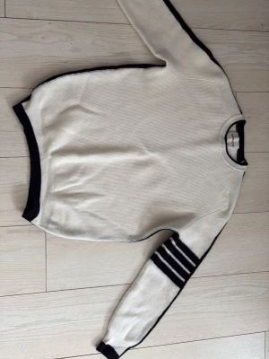 Vit stickad tröja med svarta detaljer - Säljer en stilren vit stickad tröja från Marachi clothing  med svarta kanter och fyra svarta ränder på ena ärmen. Tröjan har rund halsringning och långa ärmar. Perfekt för dig som gillar minimalistisk design och vill ha något som sticker ut lite extra. Tröjan är liten i storleken så ett steg ner storleksmässigt är att rekommendera 