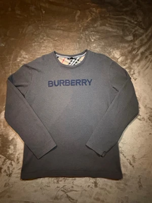 Grå långärmad tröja från Burberry - Snygg grå sweatshirt från Burberry med mörkblå broderad logga på bröstet och klassiskt rutigt mönster i halsringningen. Tröjan har rund hals och långa ärmar. Perfekt för dig som gillar stilrena och exklusiva plagg.