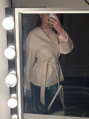 Beige omlottkappa med knytskärp - Stilren beige omlottkappa med knytskärp i midjan och slät, glansig insida. Kappan har en enkel och modern design med rak passform och långa ärmar. Perfekt för dig som gillar minimalistisk stil och vill ha en lättare kappa till vardags. Använd några fåtal gånger. Köpare står för frakt